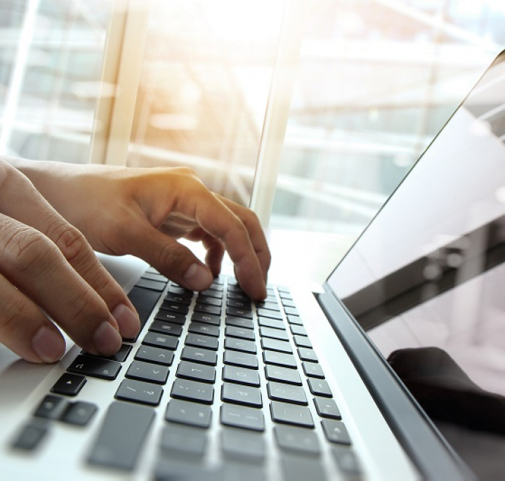 Ręce na klawiaturze otwartego laptopa, pomieszczenie w biurowcu, przez okno pada światło słoneczne