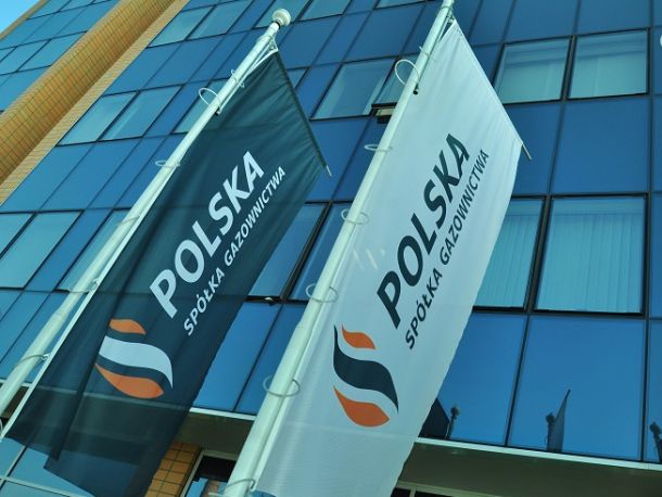 Flagi Polskiej Spółki Gazownictwa zawieszone na słupach na tle fasady szklanego biurowca