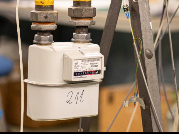 A grey gas meter connected to pipes, next to metal frame components and cables