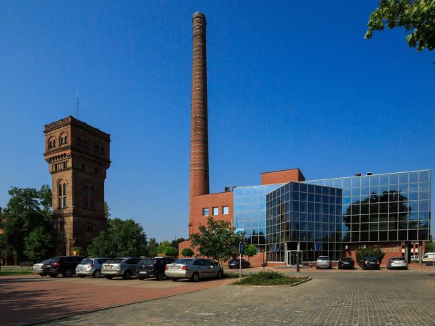 Photo showing the buildings of the former gasworks – a tower and a tall chimney, next to a modern office building, with a car park in the foreground.