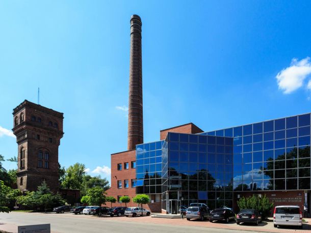 Photo showing the buildings of the former gasworks – a tower and a tall chimney, next to a modern office building, with a car park in the foreground.