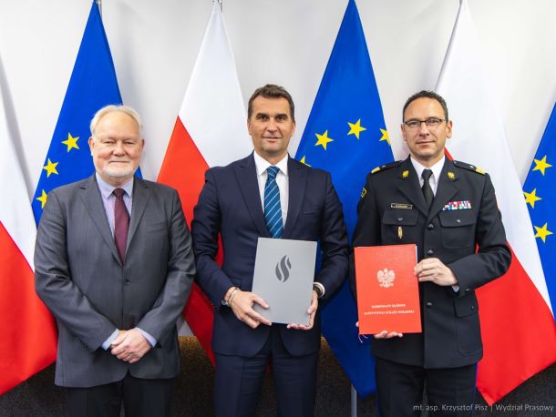 Wojciech Kruczek, Arkadiusz Kazana and Wojciech Kowalski present the signed agreement, with a wall composed of alternating Polish and European Union flags in the background