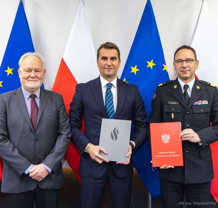 Wojciech Kruczek, Arkadiusz Kazana and Wojciech Kowalski present the signed agreement, with a wall composed of alternating Polish and European Union flags in the background
