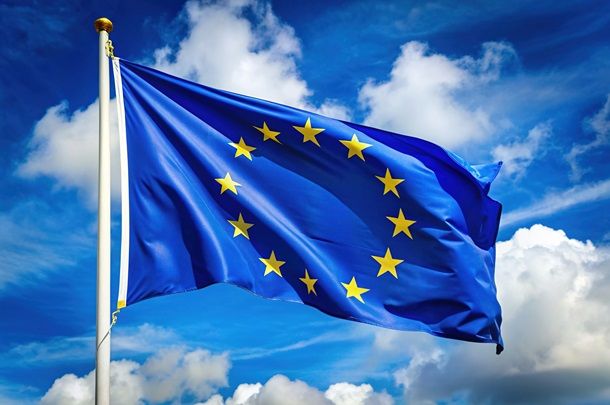 Flaga unii europejskiej wisząca na maszcie, w tle niebieskie niebo z chmurami