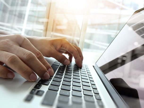 Ręce na klawiaturze otwartego laptopa, pomieszczenie w biurowcu, przez okno pada światło słoneczne